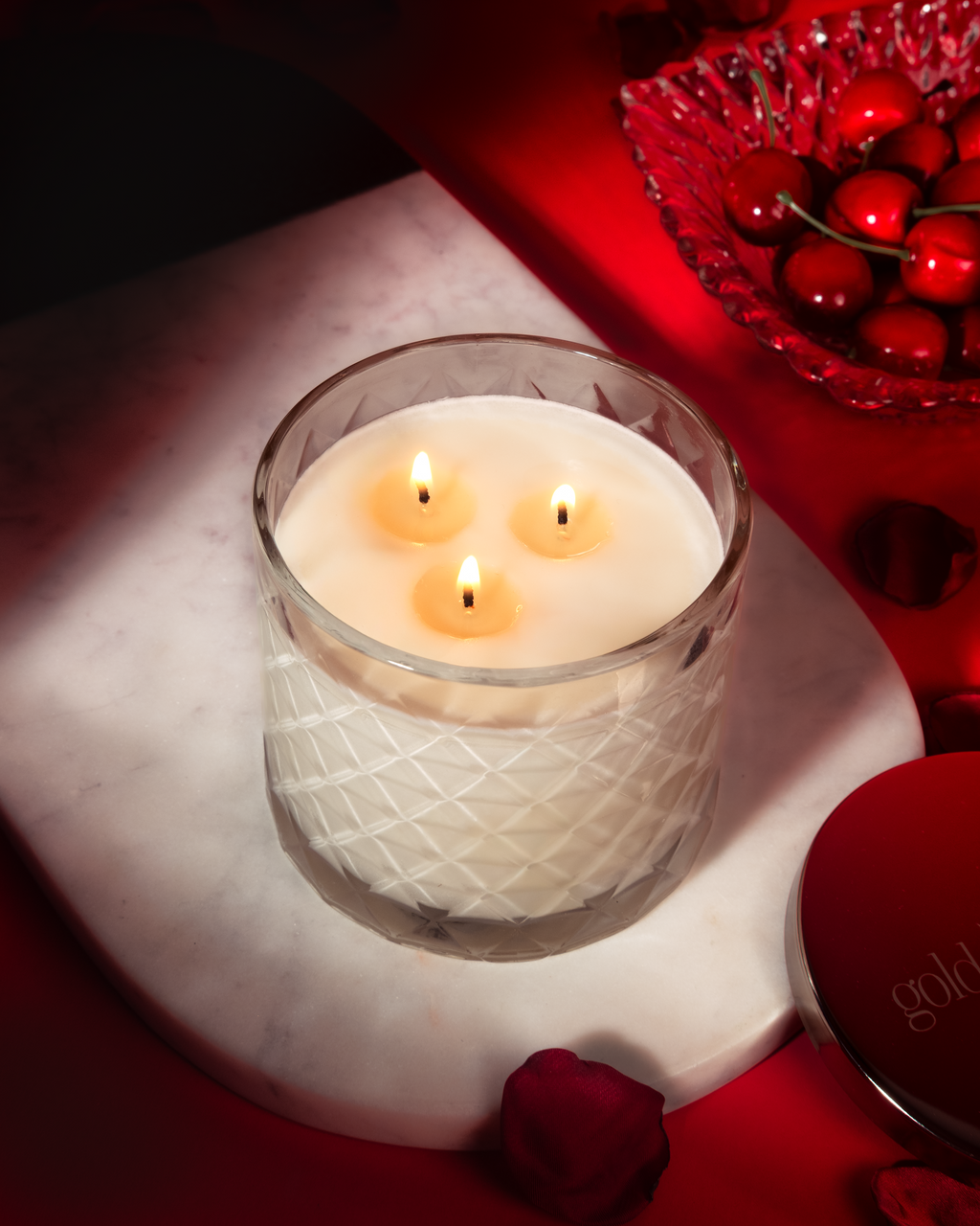 Three-wick candle in a glass holder on a marble surface with red decorative elements.