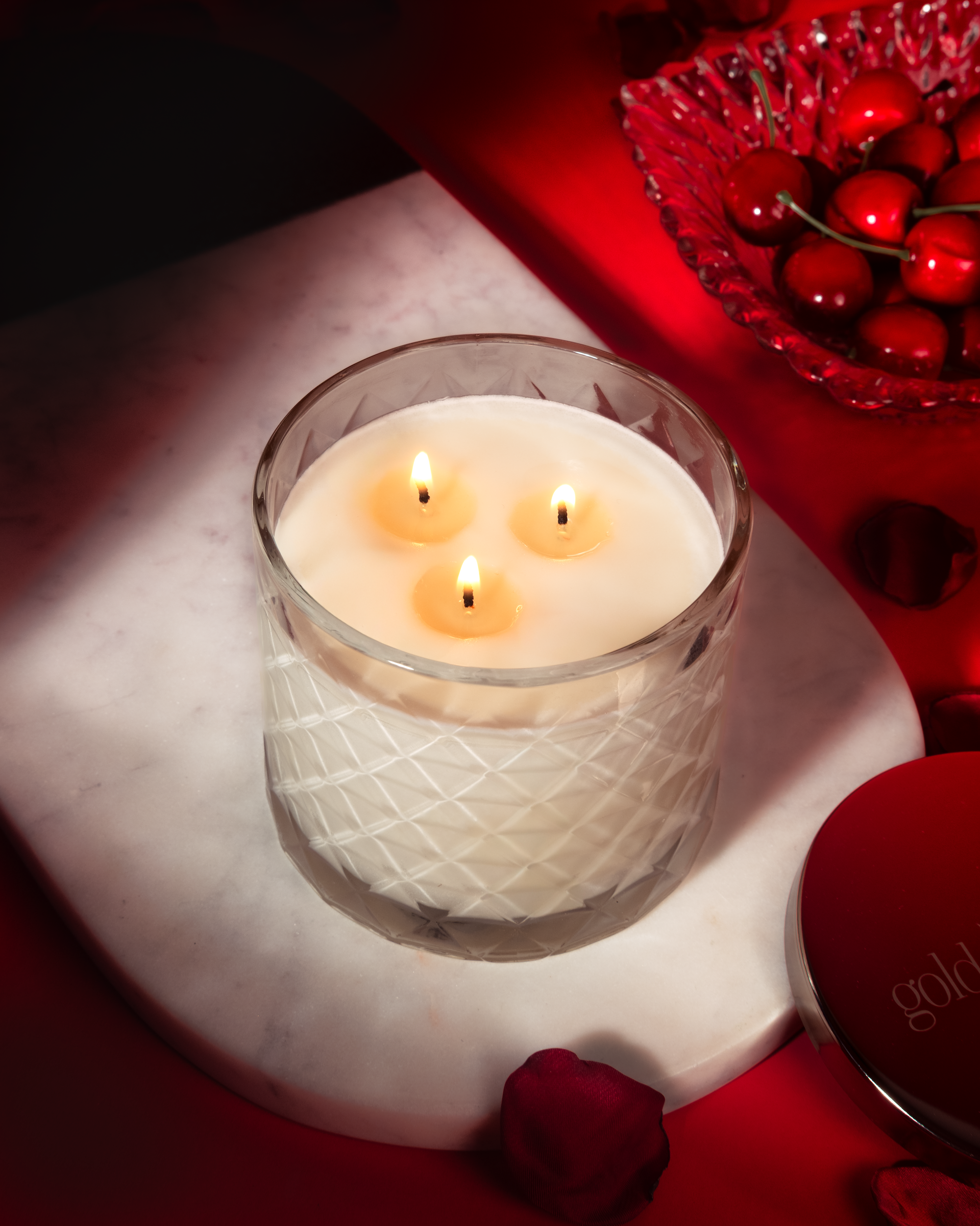 Three-wick candle in a glass holder on a marble surface with red decorative elements.