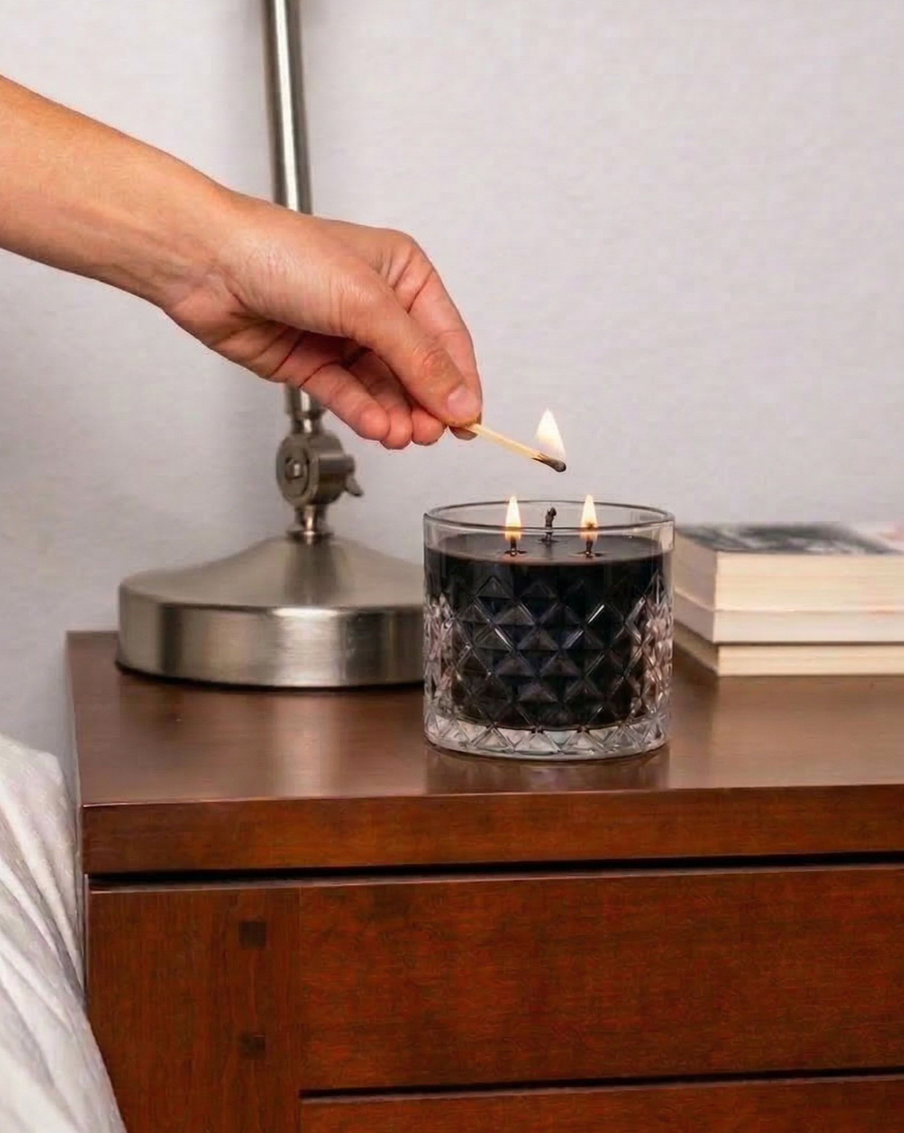Person lighting a candle on a wooden nightstand with a lamp and books in the background.